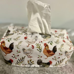 May include: A rectangular fabric tissue box cover featuring a white background with a repeating pattern of brown and white chickens, green leafy sprigs, and small red berries. A single white tissue is pulled through the top opening of the cover.
