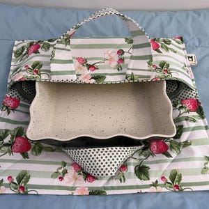 May include: A rectangular casserole dish with a cream-coloured speckled interior and dark exterior, nestled inside a quilted fabric carrier featuring a pattern of green stripes, red strawberries, and pink flowers with a green polka dot handle.