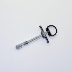 May include: A clear plastic dental syringe with a black handle and a circular thumb ring. The barrel features printed measurement markings and the text VERSA. The device includes a metal tip attachment and a black plunger mechanism for control.
