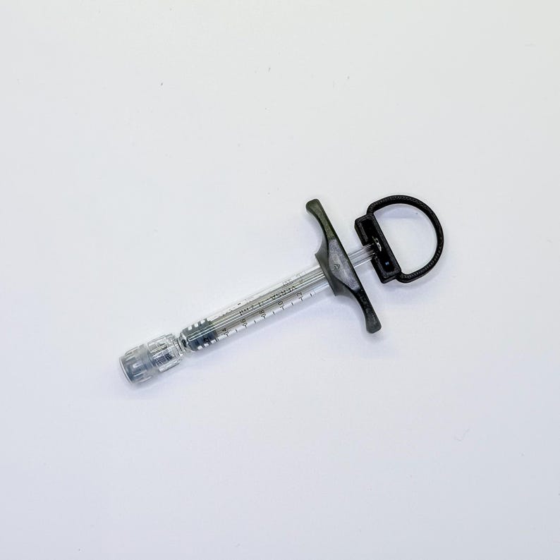 May include: A clear plastic dental syringe with a black handle and a circular thumb ring. The barrel features printed measurement markings and the text VERSA. The device includes a metal tip attachment and a black plunger mechanism for control.