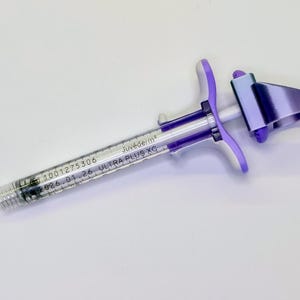 May include: A clear medical syringe with a purple plastic plunger handle and a metallic purple finger grip. The barrel is printed with the text Juv&eacute;derm ULTRA PLUS XC, the number 1001275306, and the expiration date 2026.01.26 in black ink.