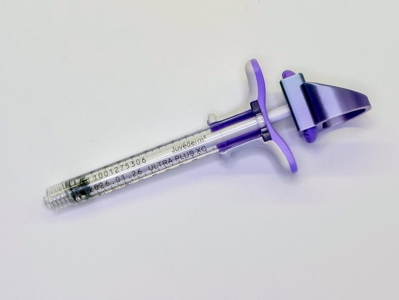 May include: A clear medical syringe with a purple plastic plunger handle and a metallic purple finger grip. The barrel is printed with the text Juv&eacute;derm ULTRA PLUS XC, the number 1001275306, and the expiration date 2026.01.26 in black ink.