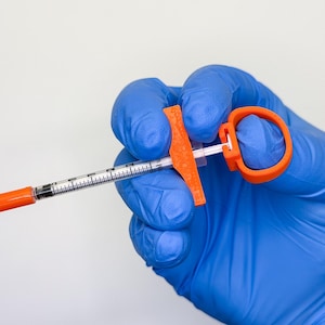 Könnte beinhalten: Eine durchsichtige Insulin-Kunststoffspritze mit orangefarbener Nadelkappe und orangefarbenem Kolbengriff. Der Spritzenzylinder weist schwarze Messmarkierungen auf. Das Gerät wird von einer Hand mit blauem Nitrilhandschuh gehalten.