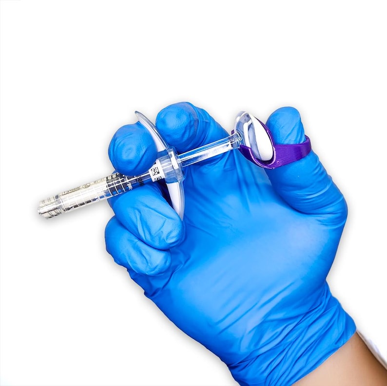 May include: A clear plastic syringe with a graduated barrel and a white plunger, featuring a purple ergonomic finger grip attachment secured to the plunger end, designed to assist with the manual operation of the syringe mechanism.