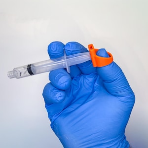 May include: A clear plastic syringe with black measurement markings on the barrel, featuring a bright orange finger ring attachment secured to the plunger end, designed to assist with grip and control during the operation of the medical device.