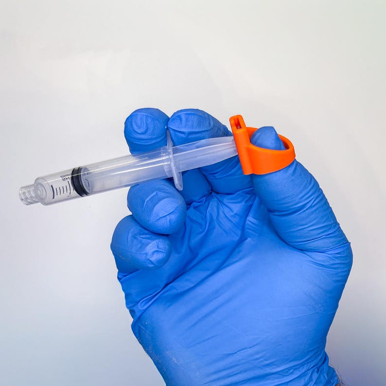 May include: A clear plastic syringe with black measurement markings on the barrel, featuring a bright orange finger ring attachment secured to the plunger end, designed to assist with grip and control during the operation of the medical device.