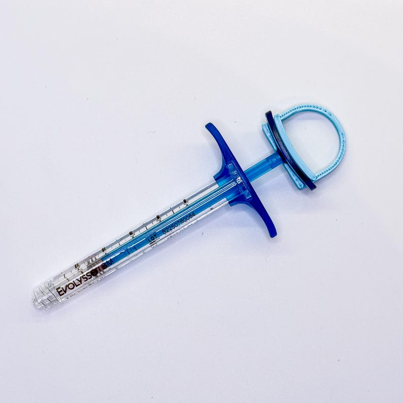 May include: A clear plastic syringe with a blue plunger and finger grip. The barrel features black measurement markings and the text EVOLYSS SM. A light blue circular thumb loop is attached to the end of the plunger rod, featuring a dark blue band.