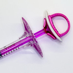 May include: A clear plastic syringe with a bright magenta plunger and a matching magenta 3D-printed finger grip attachment. The attachment features a circular loop at the end and a wide, wing-shaped flange near the center, with visible black measurement markings.