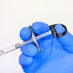 May include: A clear plastic oral syringe with a black plastic finger grip attachment and a black plastic ring pull at the plunger end. The syringe barrel features black measurement markings and the text VERSA 1 ml printed on the side.