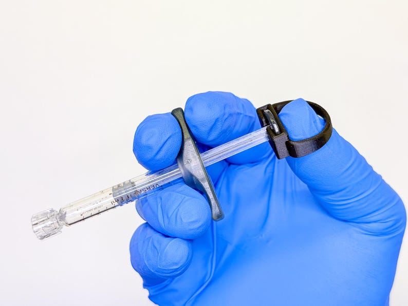 May include: A clear plastic oral syringe with a black plastic finger grip attachment and a black plastic ring pull at the plunger end. The syringe barrel features black measurement markings and the text VERSA 1 ml printed on the side.