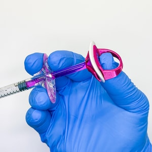 May include: A medical syringe with a purple plunger and a clear barrel containing purple liquid, fitted with a bright pink ergonomic finger-grip attachment that features a white circular top ring for enhanced control and stability during use.