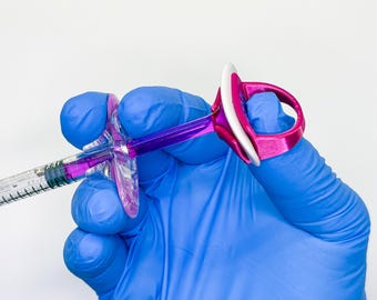 May include: A medical syringe with a purple plunger and a clear barrel containing purple liquid, fitted with a bright pink ergonomic finger-grip attachment that features a white circular top ring for enhanced control and stability during use.