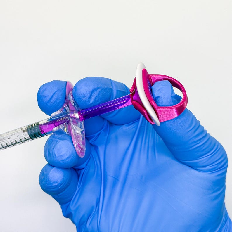 May include: A medical syringe with a purple plunger and a clear barrel containing purple liquid, fitted with a bright pink ergonomic finger-grip attachment that features a white circular top ring for enhanced control and stability during use.