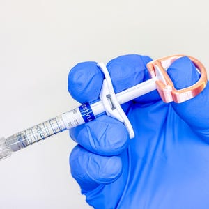 May include: A clear medical syringe with a white plunger and a copper-colored D-shaped finger grip attachment. The syringe barrel features a blue and white label with the text RESTYLANE, along with visible measurement markings and a clear cap.