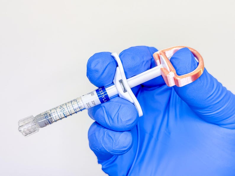 May include: A clear medical syringe with a white plunger and a copper-colored D-shaped finger grip attachment. The syringe barrel features a blue and white label with the text RESTYLANE, along with visible measurement markings and a clear cap.