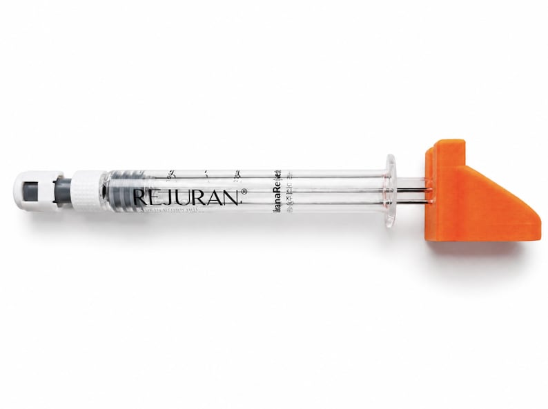 May include: A clear plastic syringe with a white cap on one end and a bright orange plastic plunger handle on the other. The barrel of the syringe features the black text REJURAN printed clearly along the side with measurement markings.