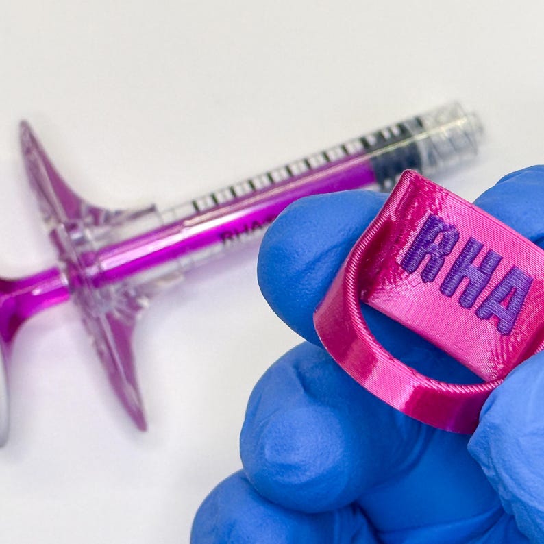 May include: A bright pink 3D-printed ring featuring the embossed letters RHA in a darker purple shade, held alongside a clear plastic syringe containing a vibrant purple liquid with the text RHA visible on its barrel.