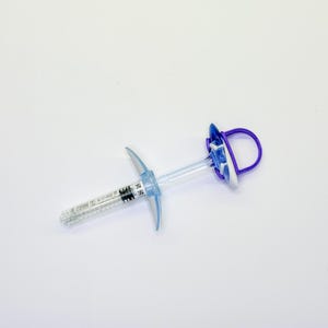 May include: A clear plastic syringe with a blue plunger handle and a purple circular finger loop. The barrel features printed black measurement markings and a small white label with a QR code and text, including the date 2024-10-19.