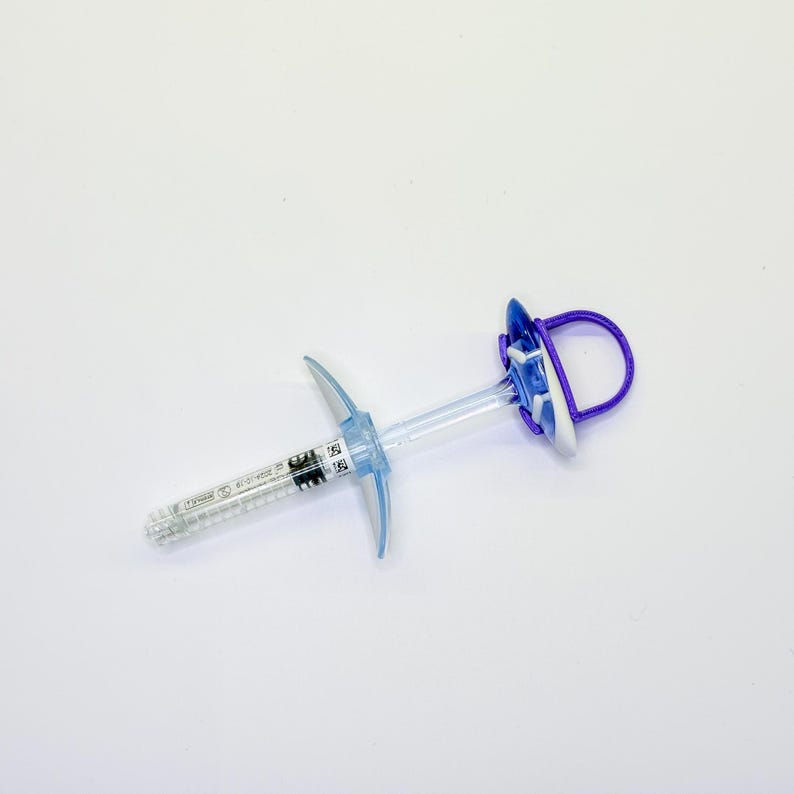 May include: A clear plastic syringe with a blue plunger handle and a purple circular finger loop. The barrel features printed black measurement markings and a small white label with a QR code and text, including the date 2024-10-19.
