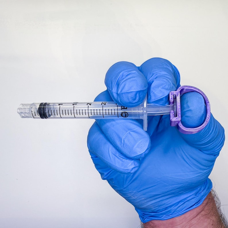 May include: A clear plastic syringe with black measurement markings and a black rubber plunger tip, featuring a purple 3D-printed ring attachment secured to the plunger end, designed to assist with grip and control during operation.