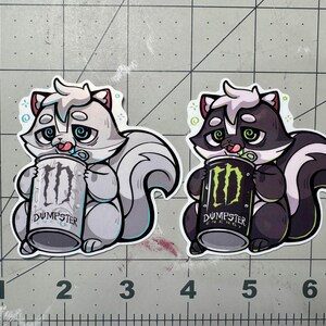 May include: Two stickers featuring cartoon skunks holding Monster Energy cans. One skunk is gray and white, the other is black and white. The cans read "Dumpster Energy" with the Monster logo.
