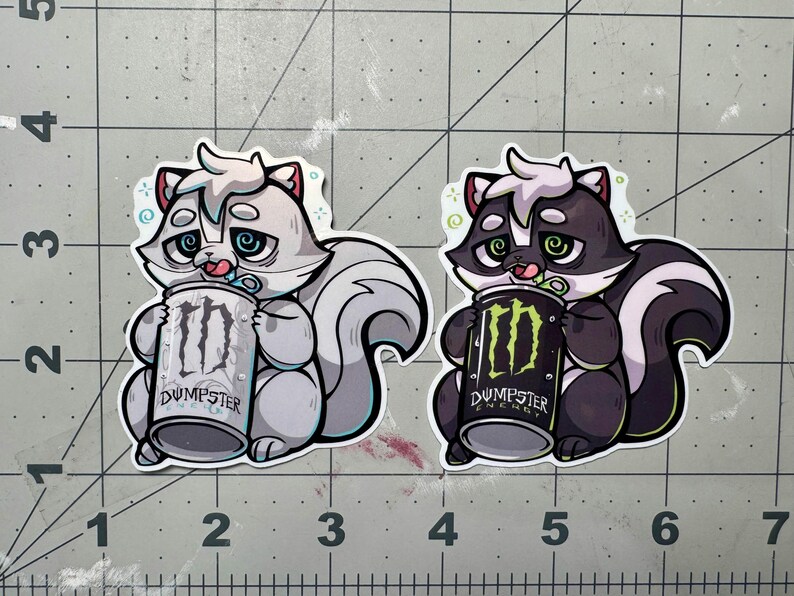 May include: Two stickers featuring cartoon skunks holding Monster Energy cans. One skunk is gray and white, the other is black and white. The cans read "Dumpster Energy" with the Monster logo.