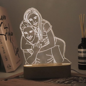 Op de afbeelding: Een witte omtrek van een koppel dat elkaar omarmt, verlicht door een warm licht, op een houten basis. Het koppel is afgebeeld in een speelse pose, waarbij de vrouw op de rug van de man zit. De basis is gemaakt van natuurlijk hout en heeft een gladde afwerking.