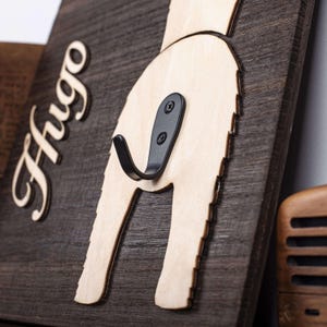 Puede incluir: Un gancho de pared de madera con forma de trasero de perro con el nombre "Hugo" tallado en la madera. El gancho es de metal negro y est&aacute; fijado a la madera con dos tornillos.