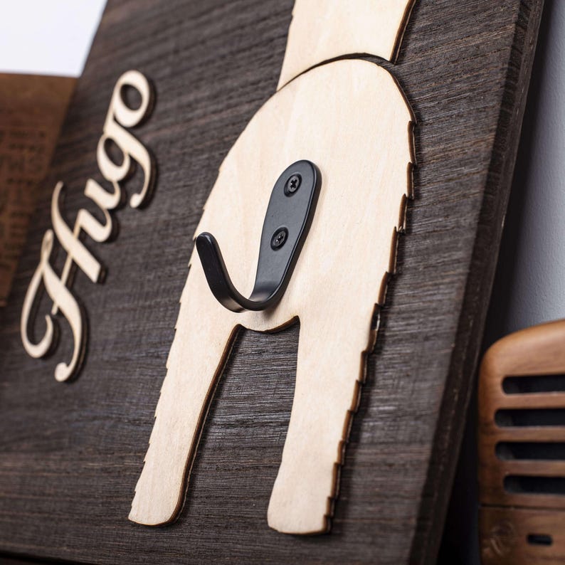 Puede incluir: Un gancho de pared de madera con forma de trasero de perro con el nombre "Hugo" tallado en la madera. El gancho es de metal negro y est&aacute; fijado a la madera con dos tornillos.