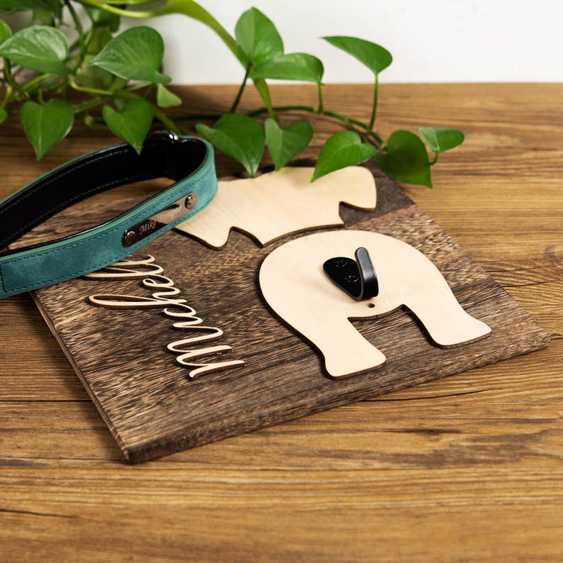 Puede incluir: Un gancho de pared de madera con forma de perro con un gancho para la correa en la parte posterior. El gancho est&aacute; pintado de marr&oacute;n y tiene el nombre "Marcella" escrito en &eacute;l. Un collar de perro verde y negro est&aacute; en primer plano.
