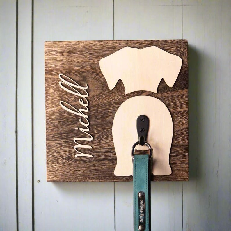 Puede incluir: Porta-correas de perro de madera con el nombre "Michelle" grabado en &eacute;l. El porta-correas tiene forma de trasero de perro con un gancho para colgar una correa. Una correa azul verdosa con una etiqueta de metal que dice "Milly Tango" cuelga del gancho.