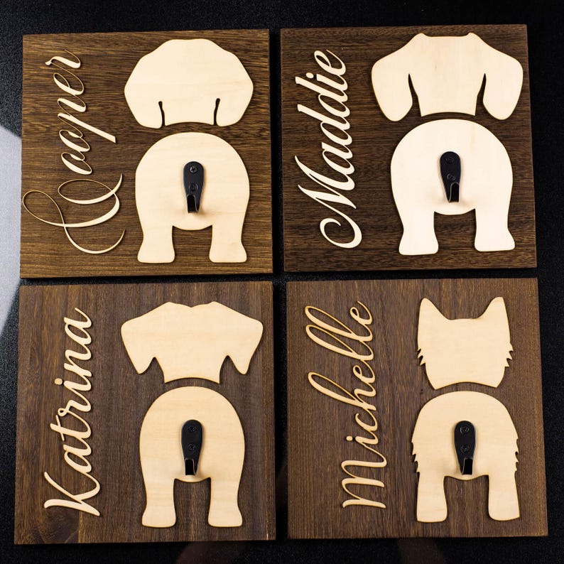 Puede incluir: Cuatro ganchos de madera con forma de trasero de perro con nombres personalizados. Los ganchos est&aacute;n pintados de marr&oacute;n y tienen un acabado de madera clara. Los nombres son "Cooper", "Maddie", "Katrina" y "Michelle".