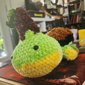 May include: A handmade crochet plush toy in the shape of a pear. The pear is primarily light green with yellow and brown accents. It has two black safety eyes and a brown stem with a green leaf. The plush is made of soft, textured yarn.