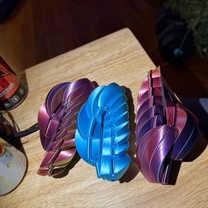 May include: Three abstract, fan-shaped sculptures in shades of blue, purple, and iridescent colours. The objects are placed on a wooden surface. The sculptures have a layered, geometric design.