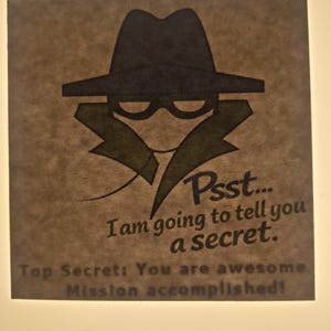 Könnte beinhalten: Braune quadratische Grafik mit einer Illustration zum Thema Spionage. Das Bild zeigt einen Fedora, eine Brille und einen Trenchcoat. Der Text lautet: „Psst... Ich werde dir ein Geheimnis verraten. Top Secret: Du bist großartig. Mission erfüllt!"