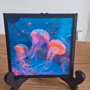 Peut inclure: Impression encadrée d'une scène sous-marine avec trois méduses. Les méduses sont représentées dans des tons de rose, d'orange et de violet, sur un fond bleu avec de petits poissons. L'impression est présentée dans un cadre noir sur un chevalet noir.