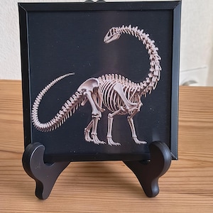 Peut inclure: Impression encadrée d'un squelette de dinosaure sur fond noir. Le squelette est détaillé et semble être un Brachiosaure. Le cadre est noir et l'impression est présentée sur un chevalet en bois.