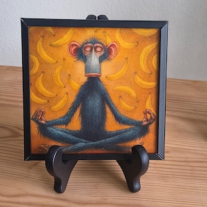 Könnte beinhalten: Gerahmtes Gemälde eines meditierenden Affen vor einem Bananen-Hintergrund. Der Affe mit roten Ohren sitzt im Lotussitz. Das Gemälde hat einen schwarzen Rahmen und steht auf einer schwarzen Staffelei. Der Hintergrund ist orange mit gelben Bananen.