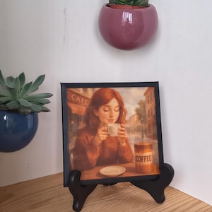 May include: A framed print of a woman enjoying coffee at a cafe, with the word "CAFE" visible. The print is displayed on a black easel. A jar of coffee is also visible. A small succulent plant is in a pink pot on the wall.