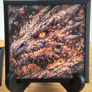 Puede incluir: Impresión enmarcada de la cabeza de un dragón, con un diseño detallado y texturizado. El ojo del dragón es de color naranja brillante, y la obra de arte tiene un tema volcánico y ardiente. El marco negro sostiene la impresión, expuesta en un caballete negro.
