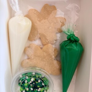 May include: A St. Patrick's Day cookie decorating kit in a white box. The kit includes three shamrock-shaped cookies, two piping bags filled with white and green icing, and a container of green sprinkles.