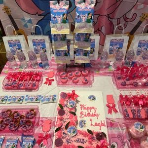 May include: A festive birthday party setup with a pink and blue theme. The table is filled with treats, including a decorated cake, cake pops, and cookies. The theme features the character Stitch, with the name "Layla" displayed.