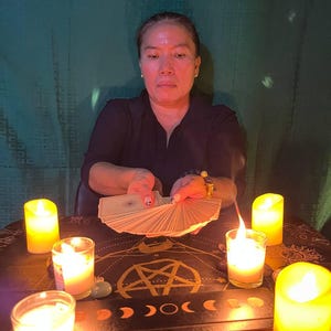 May include: A person sits at a table with a dark tablecloth and candles lit around a pentagram. They are holding a deck of tarot cards in their hands, ready to be read.