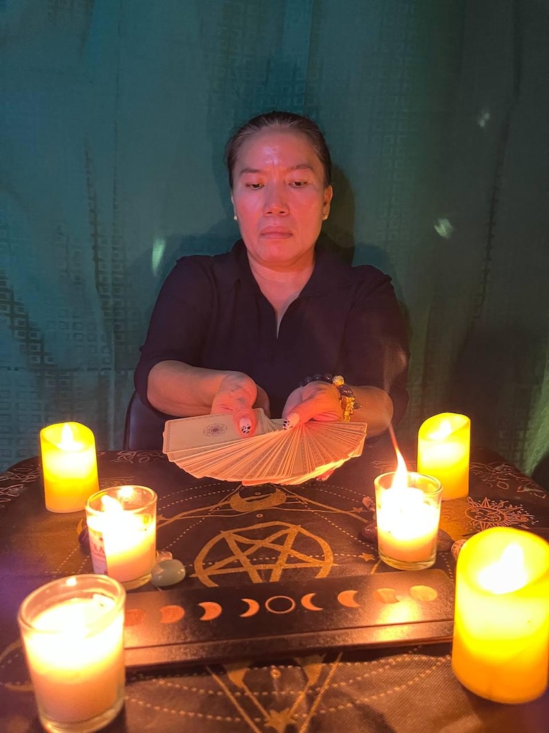 May include: A person sits at a table with a dark tablecloth and candles lit around a pentagram. They are holding a deck of tarot cards in their hands, ready to be read.