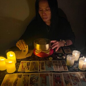 May include: A woman sits at a table with a lit candle and a bowl in front of her. She is holding a deck of tarot cards. The cards are spread out on the table, and there are several lit candles around them.