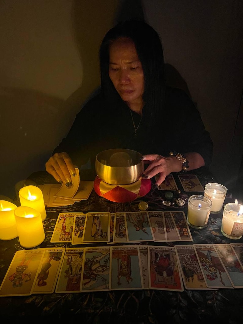 May include: A woman sits at a table with a lit candle and a bowl in front of her. She is holding a deck of tarot cards. The cards are spread out on the table, and there are several lit candles around them.