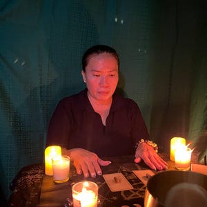 May include: A person in a black shirt sits at a table with a dark cloth featuring moon phase graphics. Three lit candles in glass holders, two small rectangular cards with stones, and a large metal singing bowl are arranged on the table surface.
