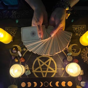 Puede incluir: Un despliegue de lectura de cartas del tarot con un pentagrama, fases lunares y velas. Una persona sostiene una baraja de cartas del tarot en sus manos.