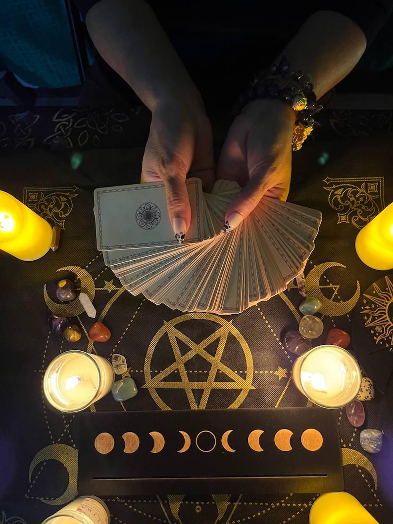 Puede incluir: Un despliegue de lectura de cartas del tarot con un pentagrama, fases lunares y velas. Una persona sostiene una baraja de cartas del tarot en sus manos.