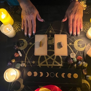 May include: A black cloth with a pentagram design, surrounded by candles, crystals, and two cards with small brown stones on them. The cloth is decorated with moons and stars.